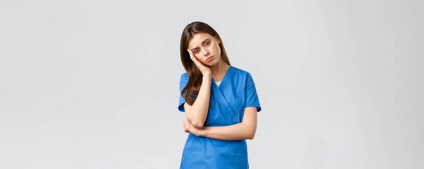 Healthcare workers, prevent virus, insurance and medicine concept. Exhausted and sleepy female nurse, doctor in scrubs, lean on palm look tired, feel fatigue from night shift at hospital - Photo, Image