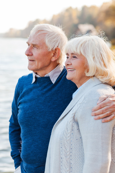 Чувствую себя в безопасности и мире. Счастливая старшая пара, стоящая вместе на набережной и улыбающаяся
 - Фото, изображение