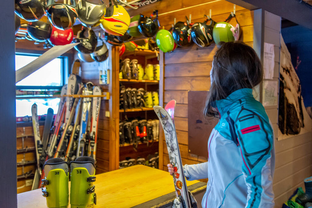 Women with brown hair renting ski equipment, waiting for her shoes and helmet, standing inside a building, lots of different helmets hanging on the roof, rear view, horizontal - Photo, Image