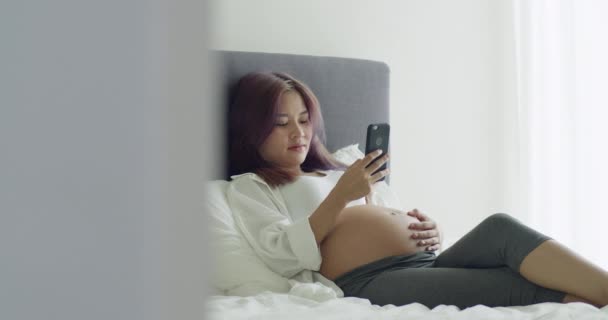 Vrolijke jonge Aziatische zwangere vrouw met behulp van smartphone terwijl liggend op een bed in de slaapkamer thuis. - Video