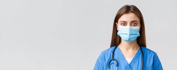 Covid-19, het voorkomen van virus, gezondheid, gezondheidswerkers en quarantaine concept. close-up jonge vrouwelijke verpleegkundige of arts in blauwe scrubs en medisch masker kijken serieus op camera - Foto, afbeelding