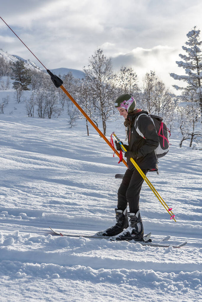Mujer esquiadora con ropa de esquí negra y mochila mirando hacia abajo, sonriendo mientras usa un telesilla, vista lateral, vertical - Foto, imagen