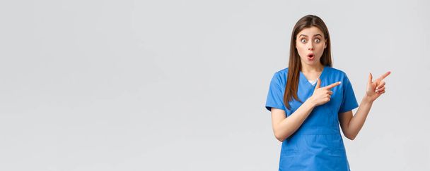 Healthcare workers, prevent virus, insurance and medicine concept. Surprised nurse or doctor in blue scrubs listen to interesting news, pointing fingers right, gasping shocked at camera - Photo, Image