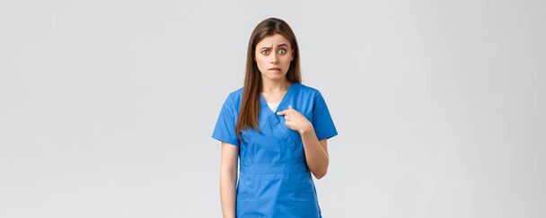 Медицинские работники, предотвратить вирус, страхование и медицина концепции. Нервная и растерянная медсестра, доктор в синей форме, кусающая губы с тревогой, указывающая на себя, сбитая с толку ожидающая ответа
 - Фото, изображение