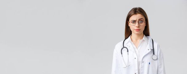 Healthcare workers, medicine, insurance and covid-19 pandemic concept. Skeptical and confused female doctor in white scrubs, medical suit and glasses, raise eyebrow judgemental, smirk displeased - Foto, afbeelding