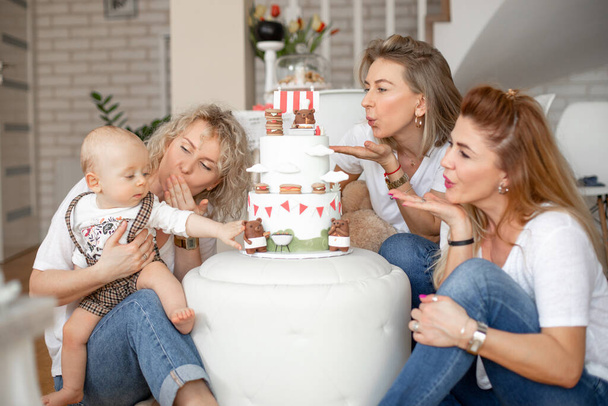 Three attractive middle-aged and young women blowing kisses to little one-year-old boy touching bear on birthday cake. - Photo, Image