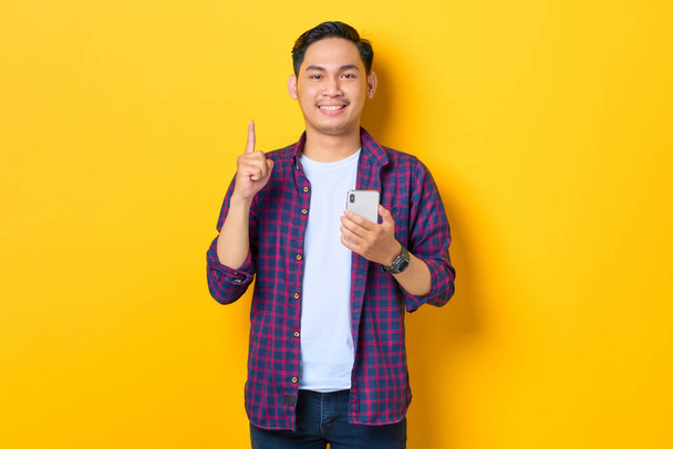 Fröhlicher junger asiatischer Mann im karierten Hemd, der sein Handy in der Hand hält und mit dem Finger nach oben zeigt und geniale Lösungen auf gelbem Hintergrund schafft - Foto, Bild