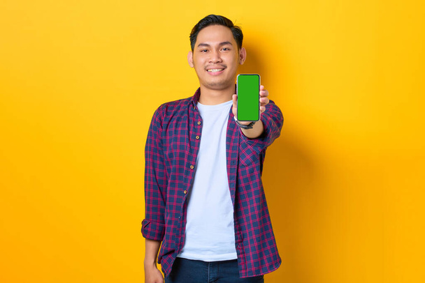 Smiling young Asian man in plaid shirt showing blank screen smartphone isolated on yellow background - Foto, imagen