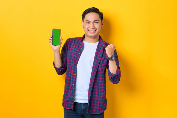 Excited young Asian man in plaid shirt showing blank screen smartphone and doing winner gesture isolated on yellow background - Zdjęcie, obraz