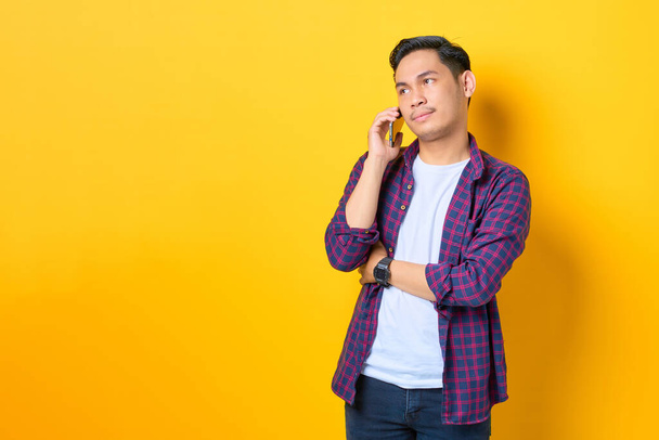 Confundido joven asiático con camisa a cuadros hablando en el teléfono móvil y mirando hacia otro lado en el espacio de copia aislado en el fondo amarillo - Foto, imagen