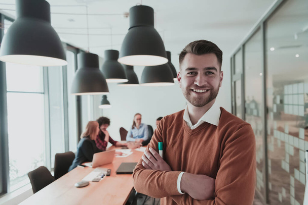 Portrait of happy millennial male business owner in modern office. Businessman wearing glasses, smiling and looking at camera. Busy diverse team working in background. Leadership concept. Head shot. - 写真・画像