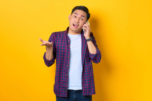 Confused young Asian man in plaid shirt talking on mobile phone isolated on yellow background - 写真・画像