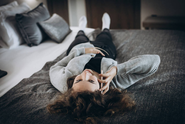 Happy smiling teenager girl talking by phone lying on her back on bed - 写真・画像
