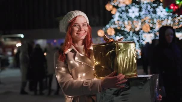 портрет счастливой молодой женщины с подарками в руках гуляет в ночном парке на фоне городской елки, улыбается и смотрит в камеру - Кадры, видео