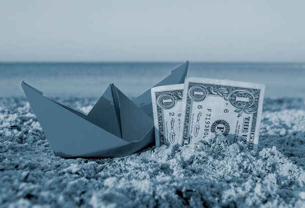 Barca di carta arancione e due banconote di carta dollaro mezzo sepolto nella sabbia sulla spiaggia sabbiosa - Foto, immagini