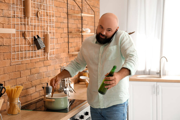 Kale man het drinken van bier en praten via de telefoon tijdens het koken in de keuken - Foto, afbeelding