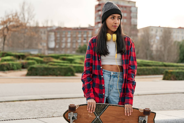 Beautiful young hipster woman posing with skateboard. Hispanic Latin woman wearing casual clothes. - 写真・画像