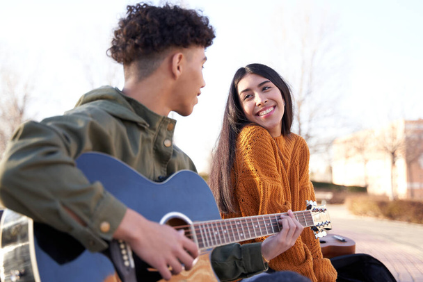 Una joven pareja latina sentada en un banco. El hombre toca una canción a la mujer que mira con amor. - Foto, imagen