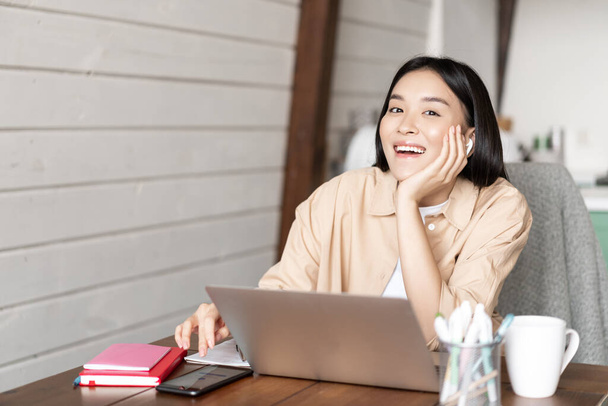 Happy asian girl listening online webinar, attend internet course classes on laptop, wearing wireless earphones and smiling, working from home - Foto, Imagem