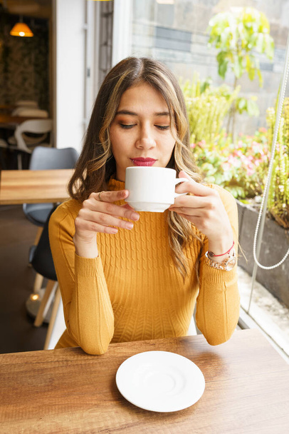 drinking and enjoying a cup of coffee a beautiful, young, latin woman with long blonde hair and makeup, restaurant interior, beauty and lifestyle with hot drink - 写真・画像
