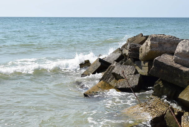 Τα κύματα της γαλαζοπράσινης θάλασσας σπάνε στις παράκτιες πέτρες και σχηματίζεται ένας πυκνός λευκός αφρός. Καθαρός μπλε ουρανός. Λευκά κύματα σπάνε σε αμμώδη παραλία μέσα από το χάσμα σε βράχους στη θάλασσα - Φωτογραφία, εικόνα