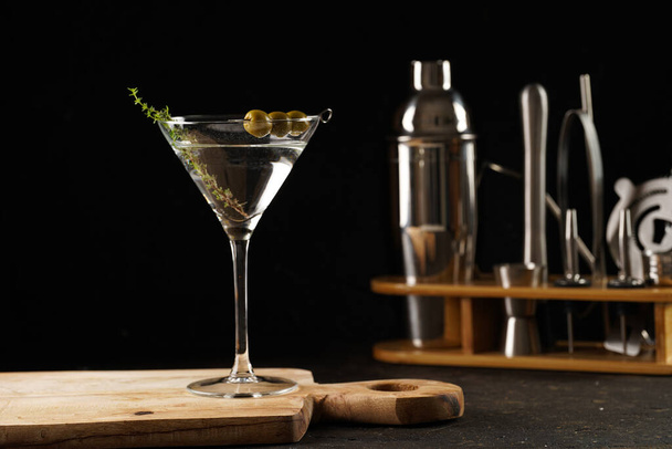 Bebida no alcohólica transparente con aceitunas y tomillo sobre tabla de madera sobre fondo negro y equipo de bar - Foto, Imagen