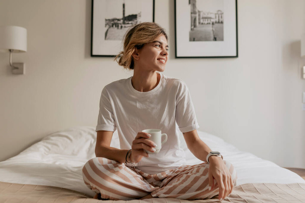 Uśmiechnięta szczęśliwa dama w piżamie siedzi rano w łóżku z kawą i patrzy przez okno w świetle słonecznym. Stylowa młoda kobieta w nowoczesnym mieszkaniu. Wysokiej jakości zdjęcie - Zdjęcie, obraz