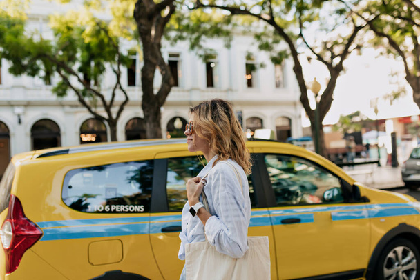 Mulher com cabelo claro ondulado em camisa azul com saco ecológico está andando na rua ensolarada luz com táxis da cidade. Menina europeia está viajando na cidade no dia quente de verão. Foto de alta qualidade - Foto, Imagem
