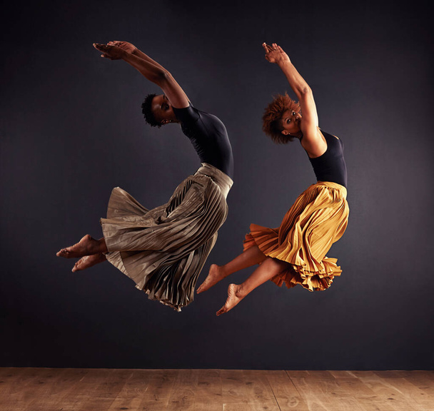 Synchronisity. Two contemporary dancers performing a synchronized leap in front of a dark background. - Zdjęcie, obraz