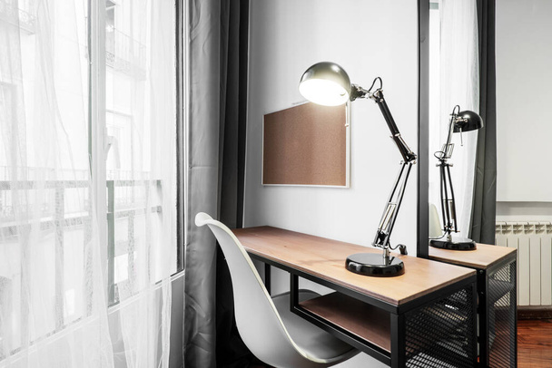 Studietafels met een witte stoel, een zwarte lamp en een spiegel aan de wand van een slaapkamer naast een raam met gordijnen en netgordijnen - Foto, afbeelding