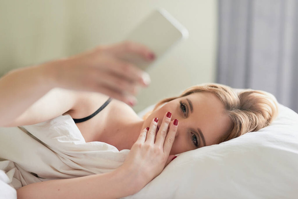 Revisando sus canales de redes sociales. Fotografía de una joven atractiva escribiendo mensajes de texto en su teléfono celular mientras yacía en su cama en casa. - Foto, Imagen