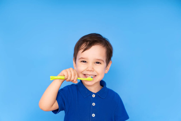 ένα μικρό όμορφο αγόρι βουρτσίζει τα δόντια του σε ένα απομονωμένο μπλε φόντο, η έννοια της στοματικής υγιεινής, ιατρική περίθαλψη. - Φωτογραφία, εικόνα