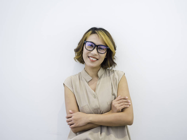 Happy smiling Asian woman short hair, trendy style in black eyeglasses and beige sleeveless shirt posing with crossed arms, looking at camera on white background. Working female portrait with glasses. - Photo, Image