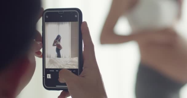 Šťastný asijský manžel pomocí smartphone pořídit fotografii těhotné ženy, zatímco stojí vedle okna v ložnici v dopoledních hodinách. - Záběry, video