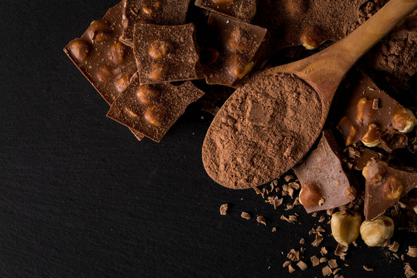 Broken chocolate nuts pieces and cocoa powder in spoon on dark stone background - Photo, image