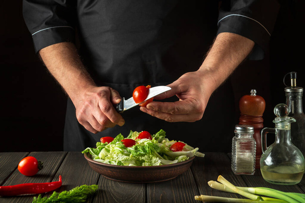 Ein professioneller Koch bereitet in der Restaurantküche einen Gemüsesalat zu. Schneiden einer frischen Tomate für einen Vitaminsalat mit einem Messer - Foto, Bild