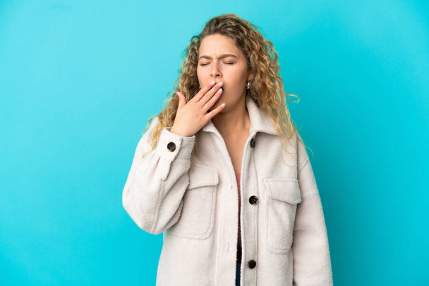 Young blonde woman isolated on blue background yawning and covering wide open mouth with hand - Photo, Image