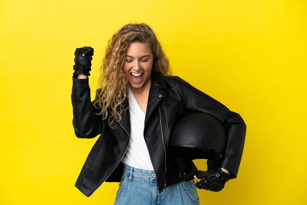 Young blonde woman with a motorcycle helmet isolated on yellow background celebrating a victory - Photo, Image