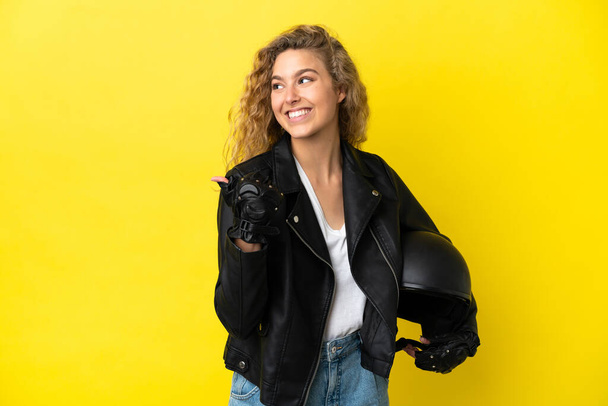 Fiatal szőke nő egy motorkerékpár sisak elszigetelt sárga háttér mutatva az oldalra, hogy bemutassa a termék - Fotó, kép