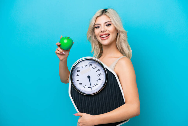 Young Russian woman isolated on blue background with weighing machine and with an apple - Photo, Image
