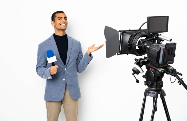 Δημοσιογράφος Αφροαμερικανός που κρατάει μικρόφωνο και μεταδίδει ειδήσεις σε απομονωμένο λευκό φόντο που απλώνει τα χέρια στο πλάι για πρόσκληση να έρθει - Φωτογραφία, εικόνα