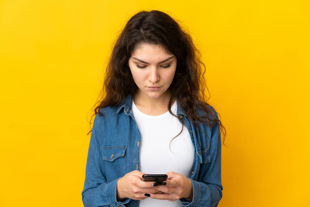 Подростковая русская девушка изолирована на желтом фоне с помощью мобильного телефона - Фото, изображение