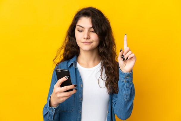 Teenager Russian girl isolated on yellow background using mobile phone and lifting finger - Zdjęcie, obraz