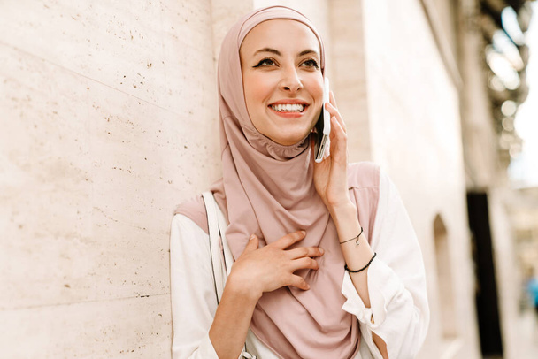 Close up of a smiling young muslim woman in hijab standing on a street using mobile phone - Фото, изображение