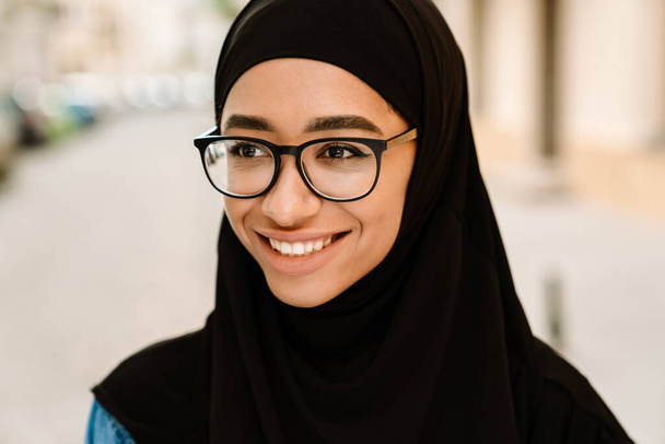 Close up of a smiling young muslim woman in hijab standing on a street - Photo, image