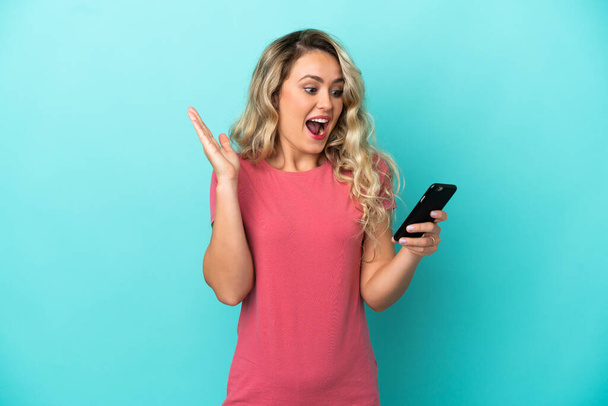 Young Brazilian woman isolated on blue background looking at the camera while using the mobile with surprised expression - Photo, Image