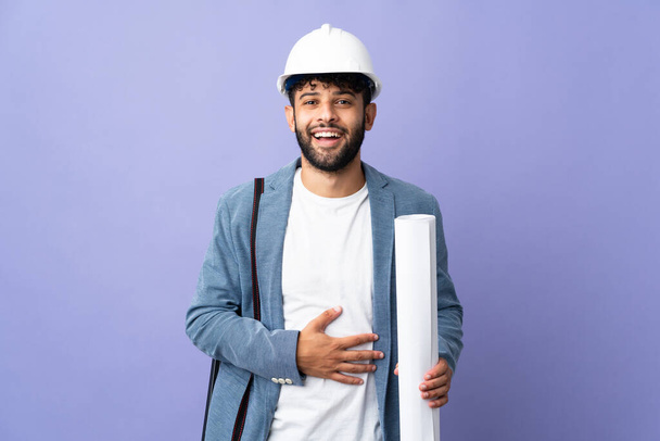 Young architect Moroccan man with helmet and holding blueprints over isolated background smiling a lot - Foto, Imagem