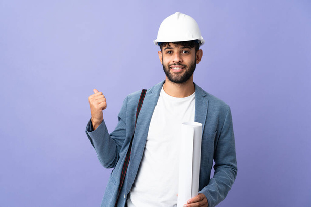 Young architect Moroccan man with helmet and holding blueprints over isolated background pointing back - Photo, Image