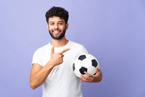 Genç Faslı adam mor arka planda, futbol topuyla izole edilmiş ve yan tarafı işaret ediyor. - Fotoğraf, Görsel