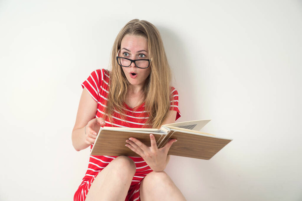 Chica rubia en un vestido de rayas rojas con gafas y un libro sobre un fondo blanco. Sorpresa.. - Foto, imagen